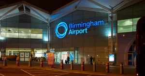 Airport terminal exterior with parked cars, traffic cones, and clear blue sky in the background.