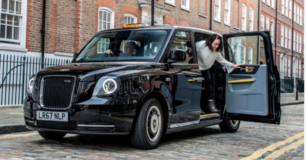 A black London-style taxi stopped on a cobblestone street as a woman steps out of the open passenger door.