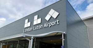 Exterior view of the London Luton Airport building with large white signage on a modern grey facade under a cloudy sky.