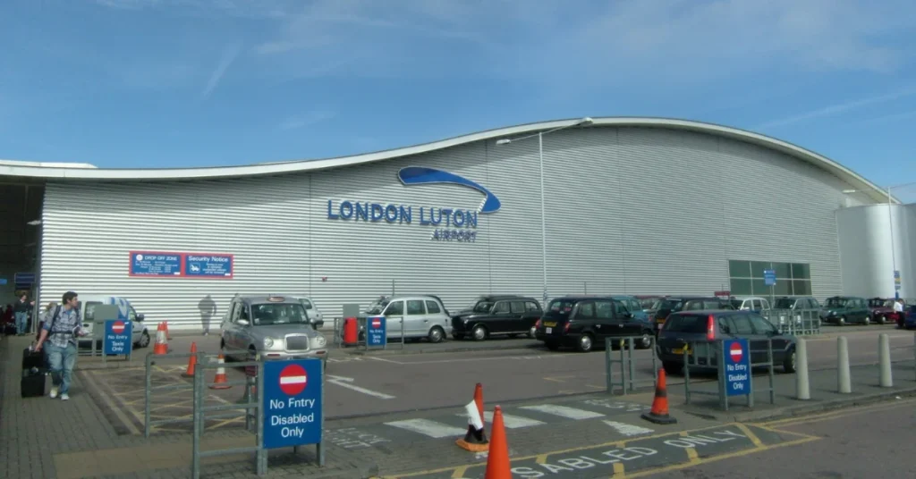 Exterior view of the London Luton Airport building with large white signage on a modern grey facade under a cloudy sky.